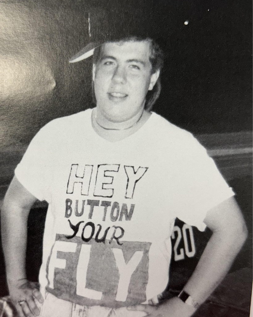 Black-and-white photograph of a smiling person standing with their hands on their hips, wearing a baseball cap and a T-shirt that reads “HEY BUTTON YOUR FLY.” The person is photographed from the waist up against a dark background.