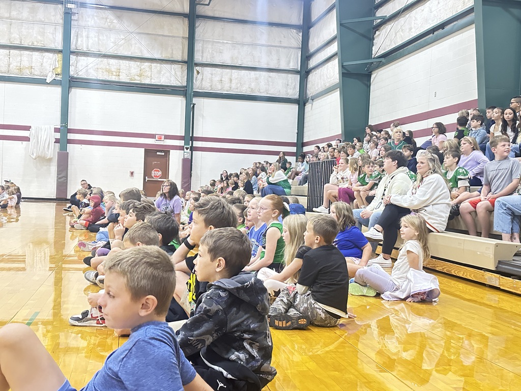 students are sitting on the floor and in the bleachers for the assembly