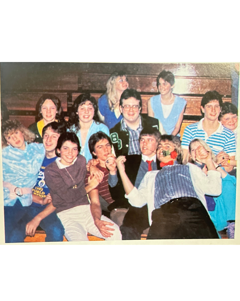 A 1986 colored old photo of high schoolers in the gym on bleachers posing for a photo and looking silly. 