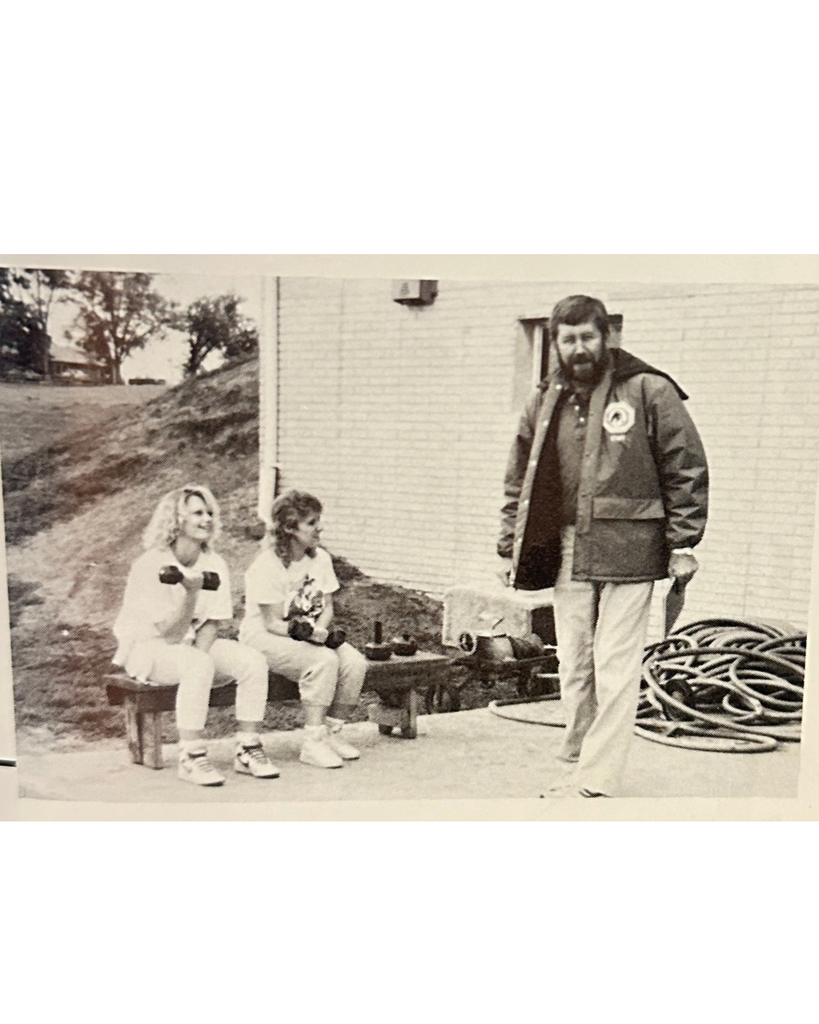An old photo from 1988 with a tall guy standing and a garden hose behind him. Two ladies on a bench holding weights outside of a school building with trees in the background.