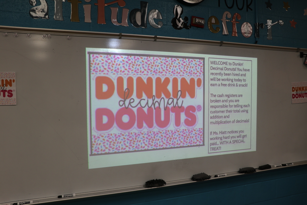 The instructions for the assignment are projected onto the board. It says Welcome to Dunkin' Decimal Donuts!nYou have recently been hired and will be working today to earn a free drink and snack. The cash registers are broked and you are responsible for telling each customer their total using addition and multiplication of decimals.