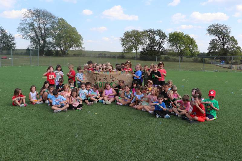 1st Graders blow bubbles while sitting on the new turf around a sign that says "Thank You Voters".