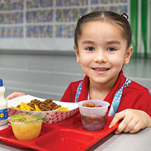 Elementary student sitting down eating her school-provided lunch on a lunch tray