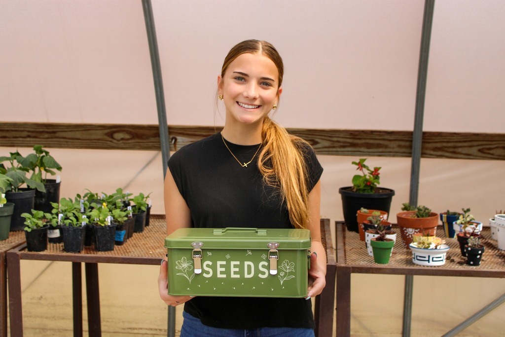 Smiling student holding a green box that says Seeds on the front. There are individually potted plants behine her