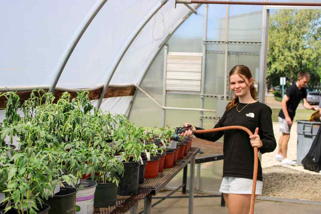 student holding hose watering individually potted plants in greenhouse