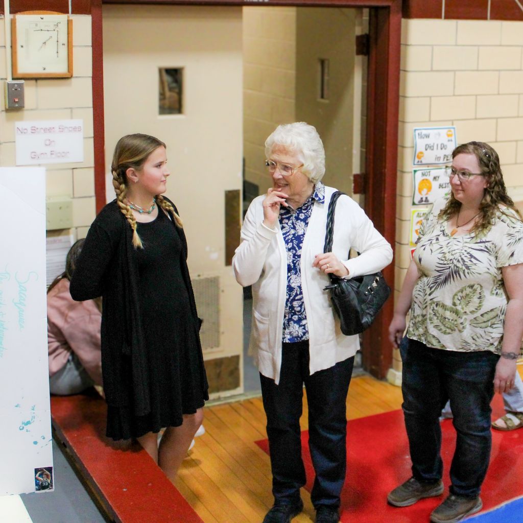 Student dressed as Sacajawea surrounded by older adults. They are presenting a display board featuring facts about the historical figue  in a school gymnasium.