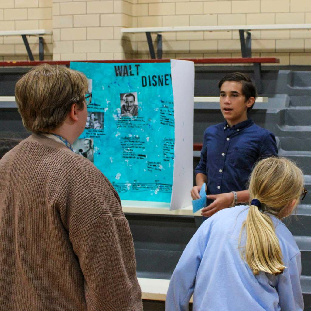 Student dressed as Walt Disney surrounded by students. They are presenting a display board featuring facts about the historical figue  in a school gymnasium.