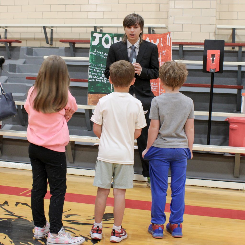 Student dressed in a suit surrounded by students. They are presenting a display board featuring facts about a historical figue  in a school gymnasium.