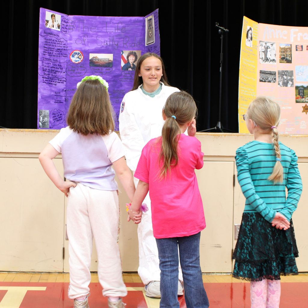 Student dressed as Sally Ride surrounded by students. They are presenting a display board featuring facts about the historical figue  in a school gymnasium.