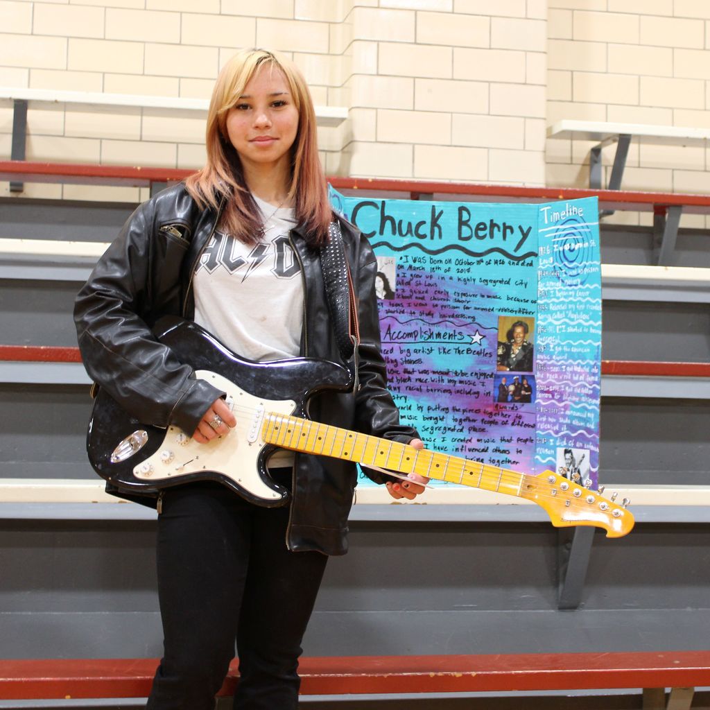 Student dressed as Chuck Berry in front of a display board featuring facts about the historical figue  in a school gymnasium.