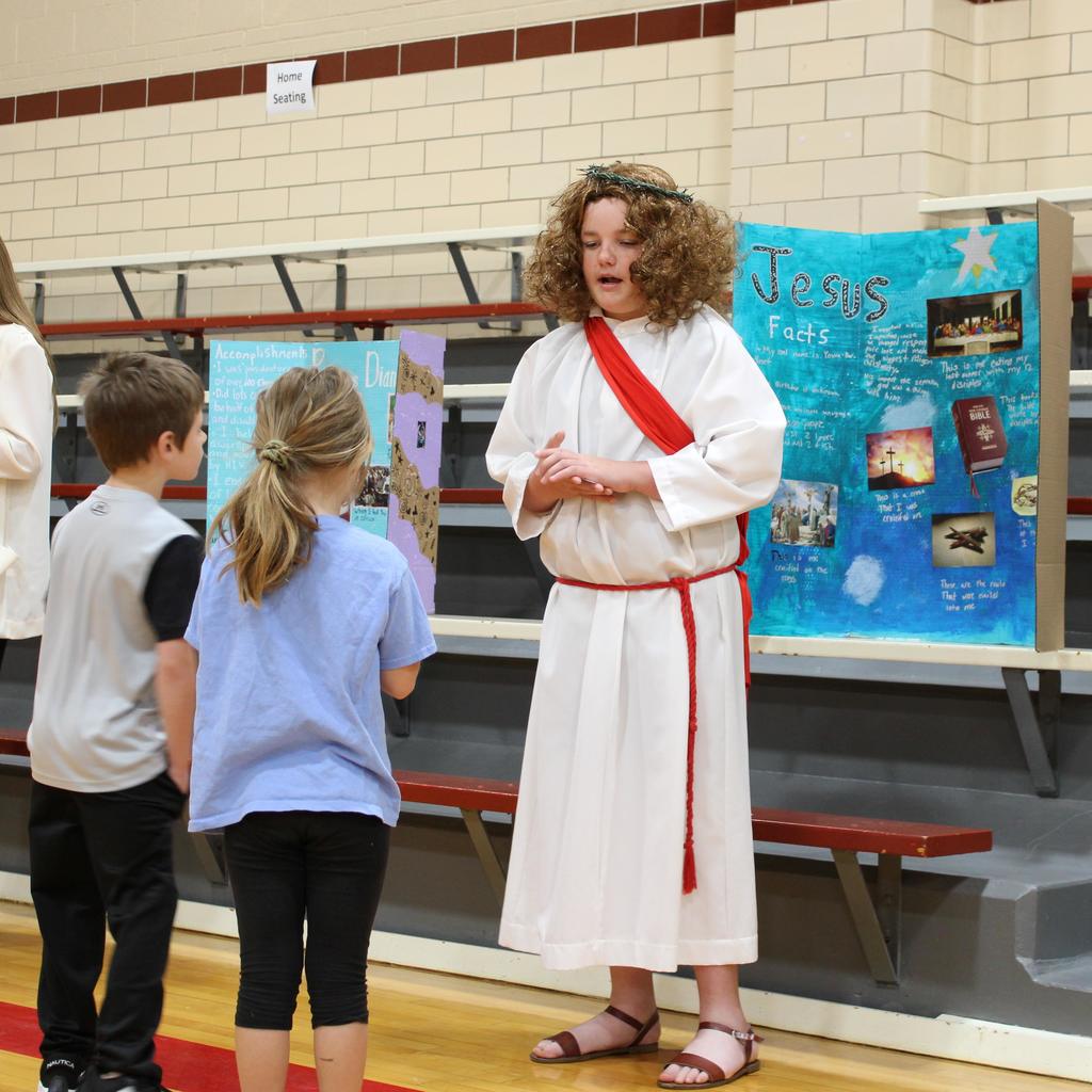 Student dressed as Jesus surrounded by students. They are presenting a display board featuring facts about the historical figue  in a school gymnasium.