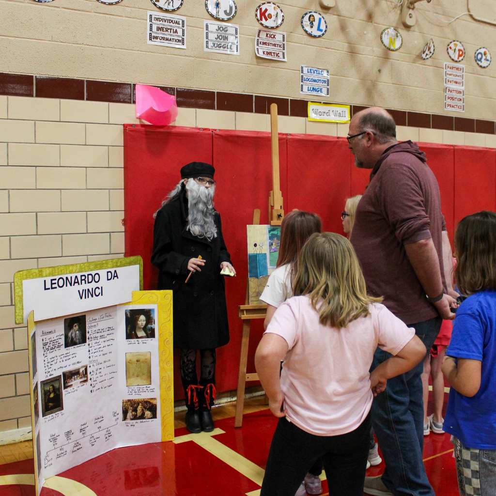 Student dressed as Leonardo da Vinci surrounded by students. They are presenting a look display board featuring the artist's famous works in a school gymnasium.