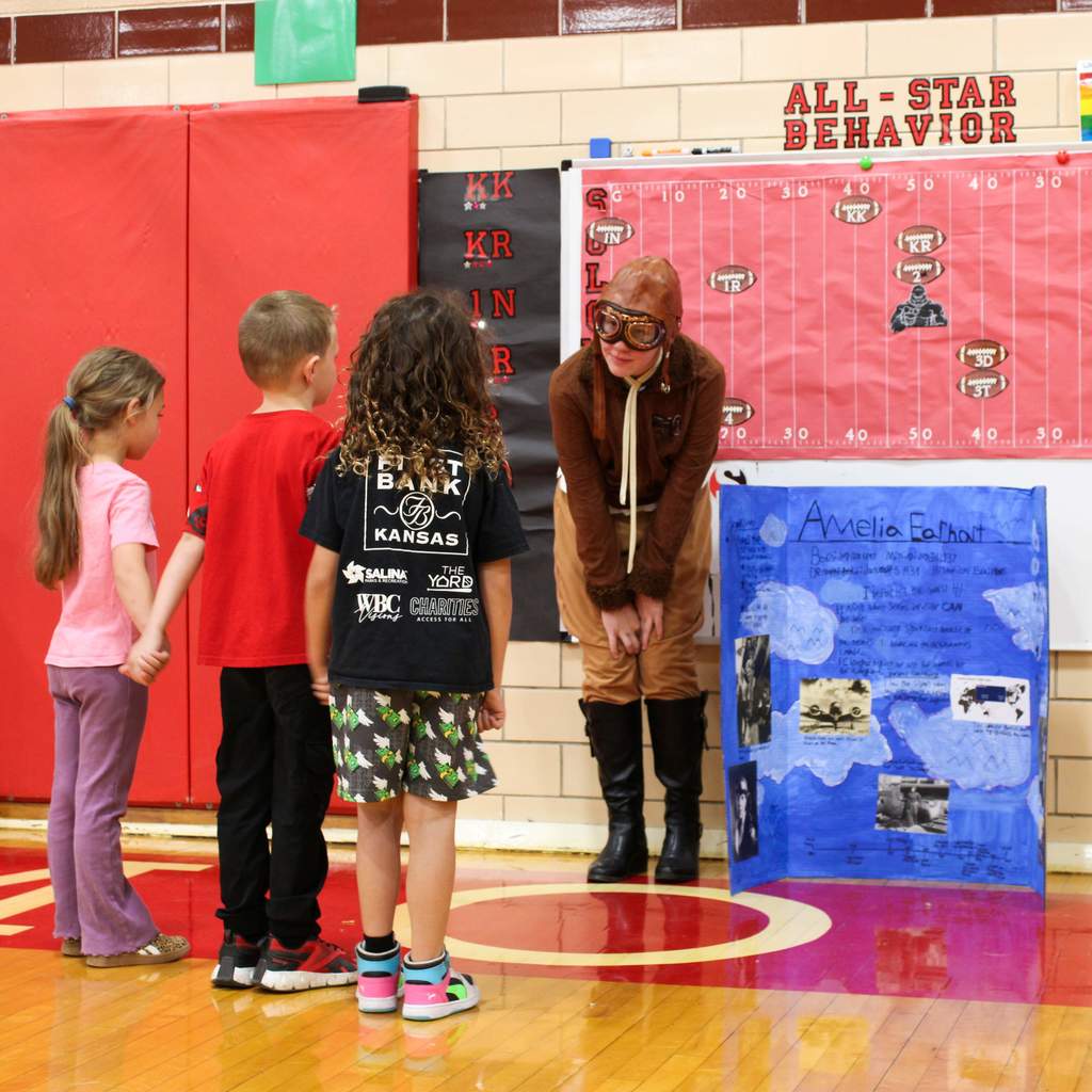 Student dressed as Amelia Earhart surrounded by students. They are presenting a display board featuring facts about the historical figue  in a school gymnasium.