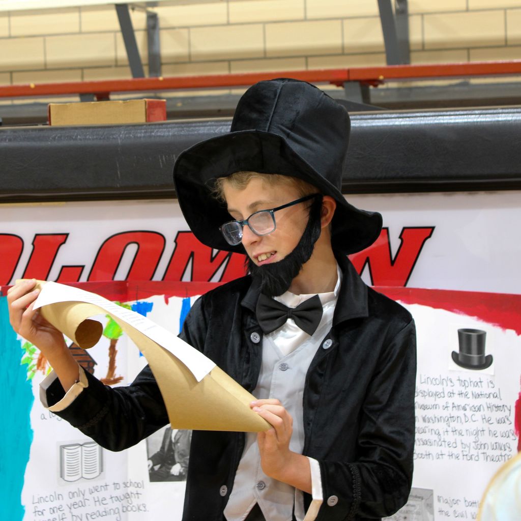 Student dressed as Abraham Lincoln. They are presenting a display board featuring facts about the historical figue  in a school gymnasium.