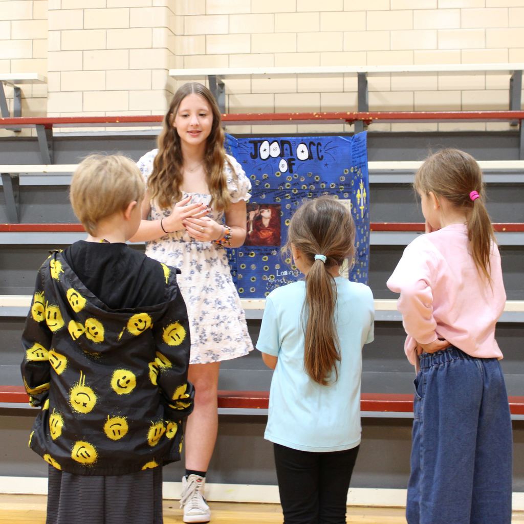 Student dressed asJoan of Arc surrounded by students. They are presenting a display board featuring facts about the historical figue  in a school gymnasium.