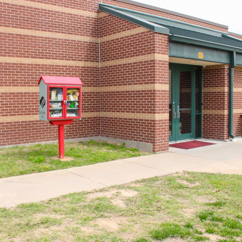 A red community sharing pantry box mounted on a post outside a brick building, stocked with various food items and canned goods.