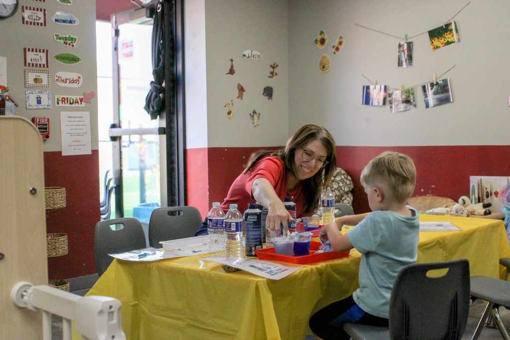 A teacher and a young boy sitting at a yellow table in a classroom, working together on a hands-on science or sensory activity involving bottles and trays.