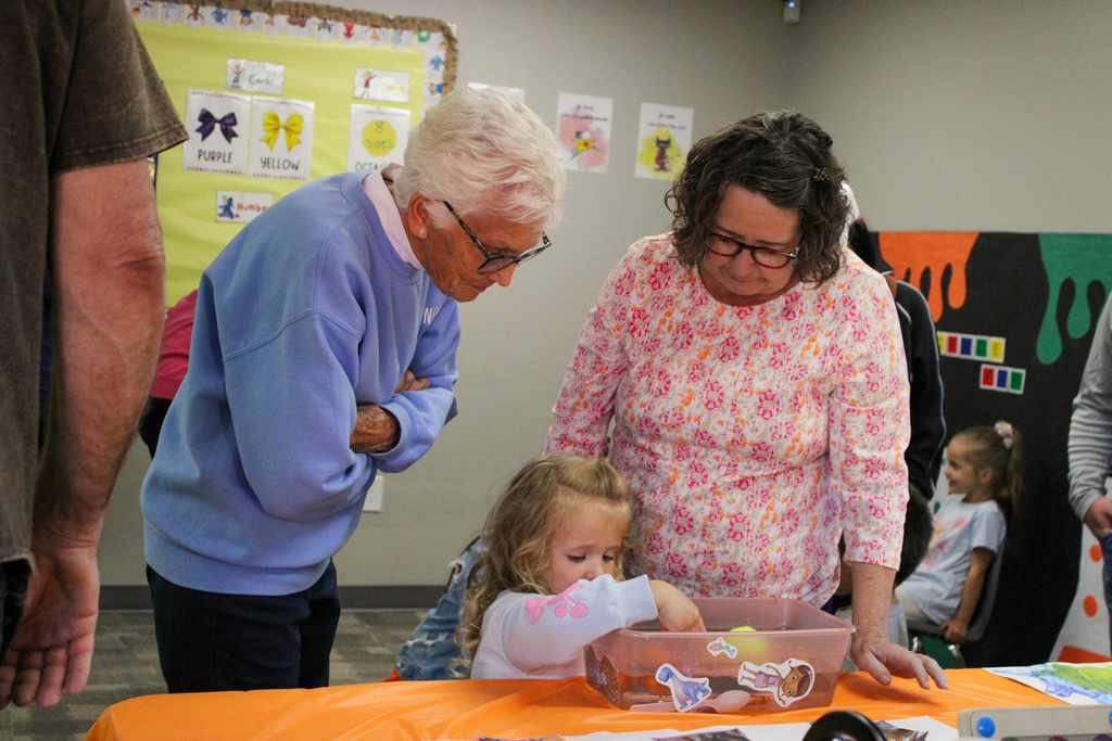 An adult mentor or teacher helping a young girl with a seated activity at a classroom table.