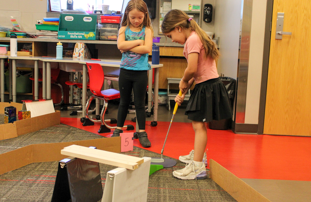 two students, one with a golf club, one on looking, mid play on their mini-golf course