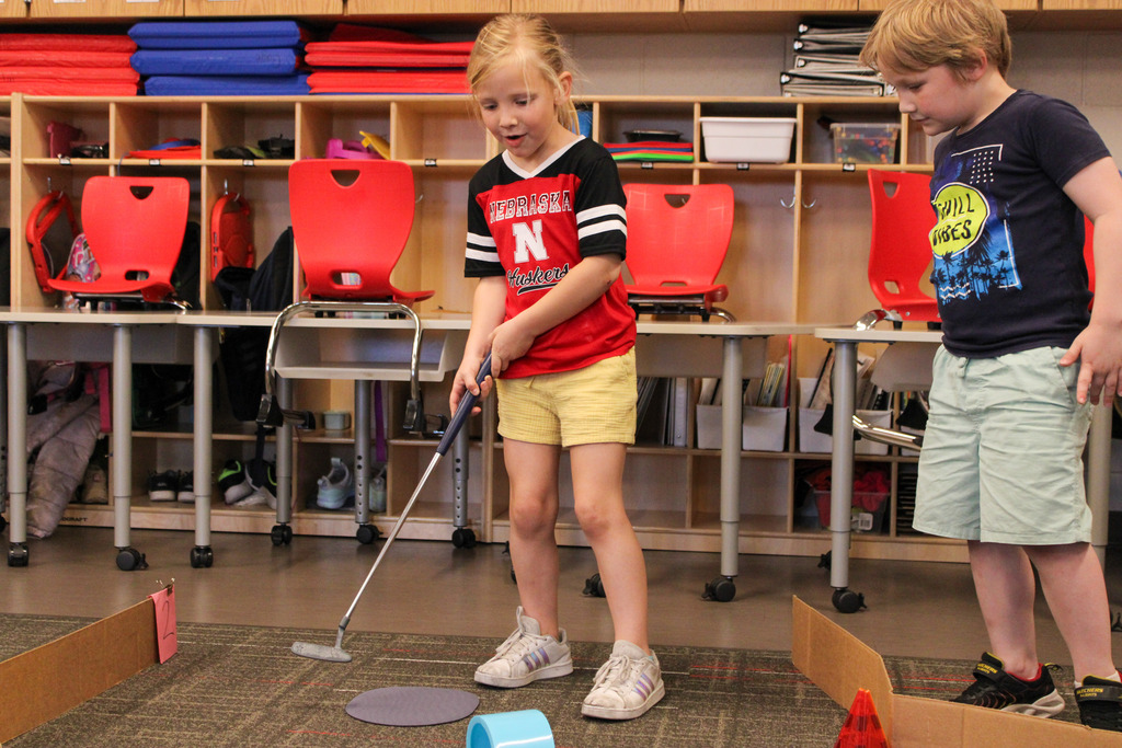 two students, one with a golf club, one on looking, mid play on their mini-golf course