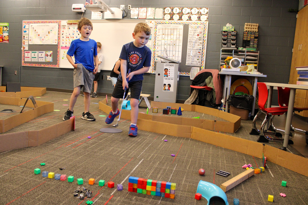 two students, one with a golf club, one on looking, mid play on their mini-golf course