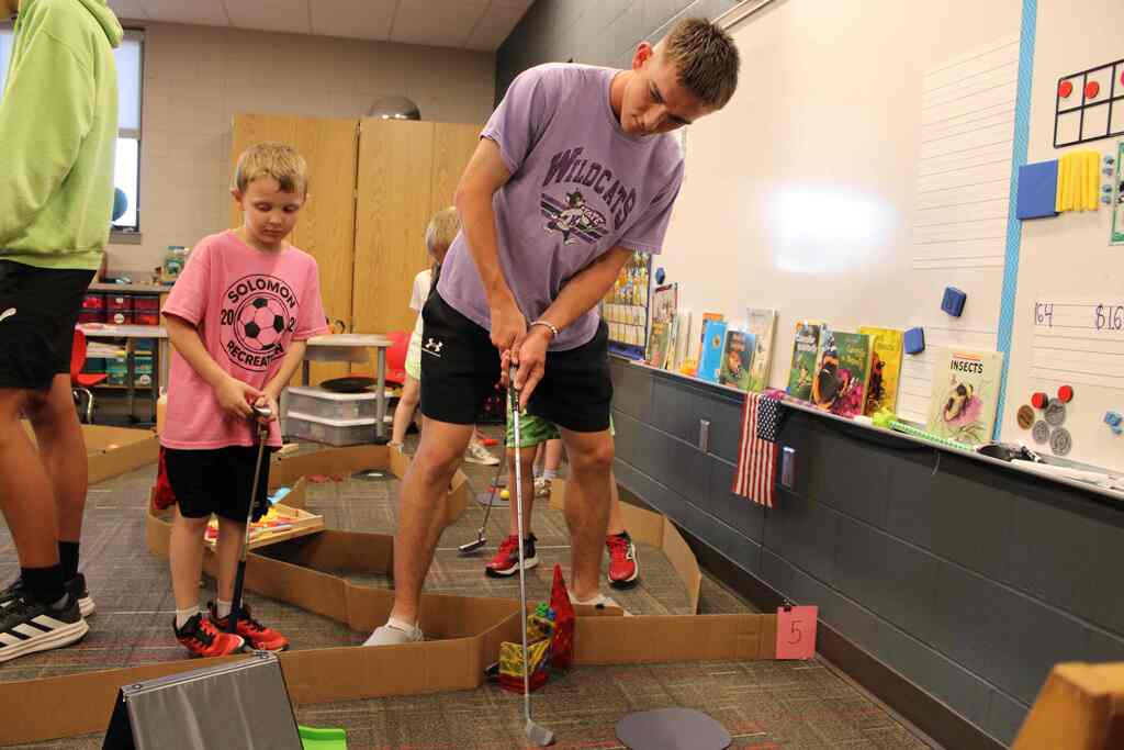 two students, the older one with a golf club, the younger one on looking, mid play on their mini-golf course