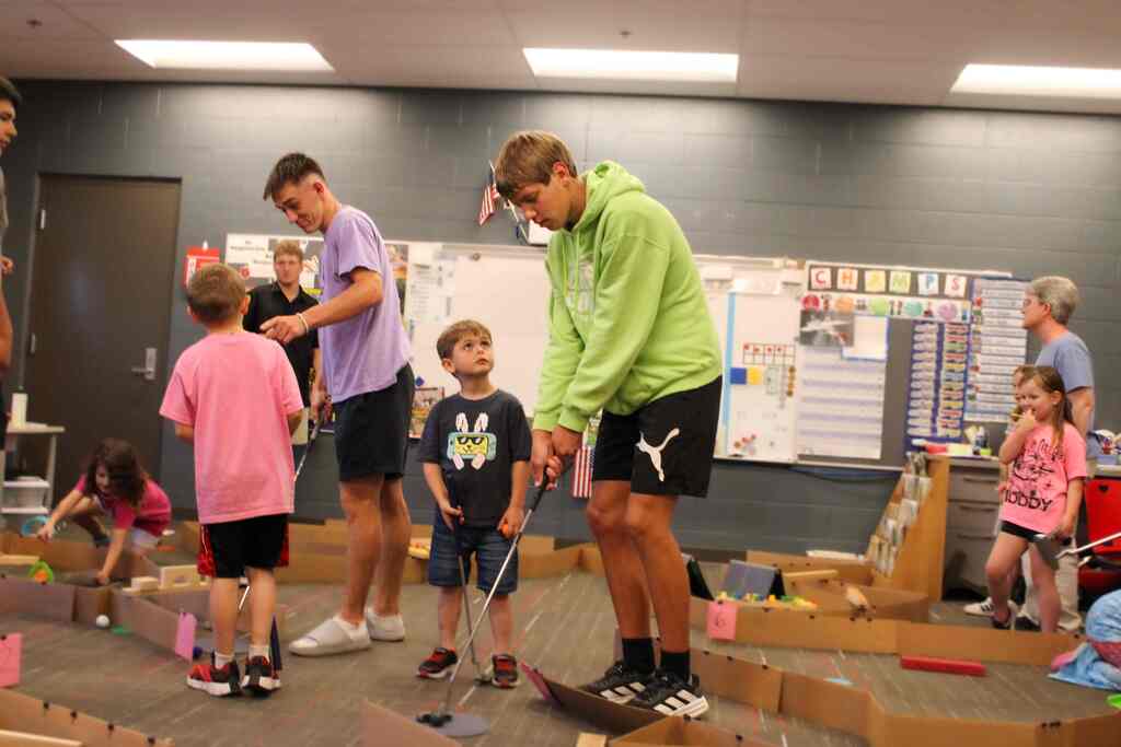 two students, the older one with a golf club, the younger one on looking, mid play on their mini-golf course