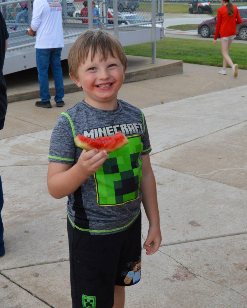 student smiling while holding a watermelon slice, red tint all along his mouth from fruit