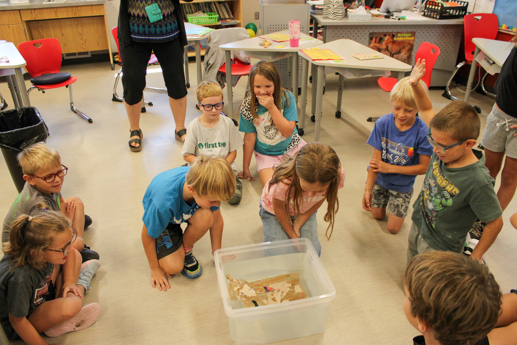 group of students sitting around a bucket of water, waiting to see if the boat on top will sink or float