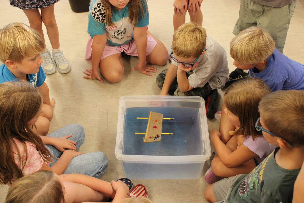 group of students sitting around a bucket of water, waiting to see if the boat on top will sink or float