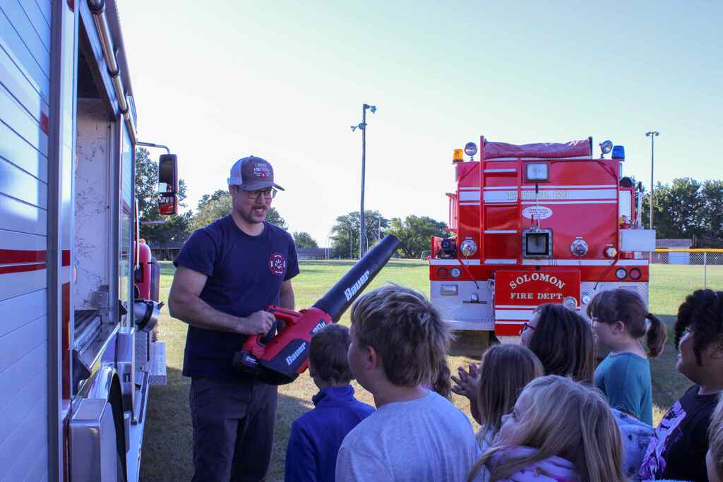 students listening to a presentation from a firefighter who is beling playful and holding a leaf blower