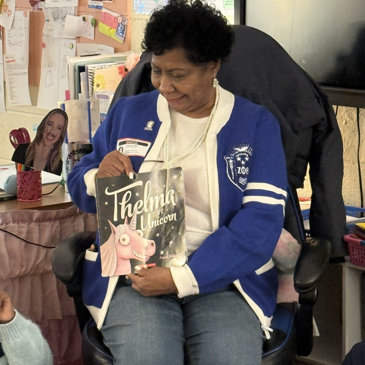 woman in blue sweater reading a book to students in a classroom