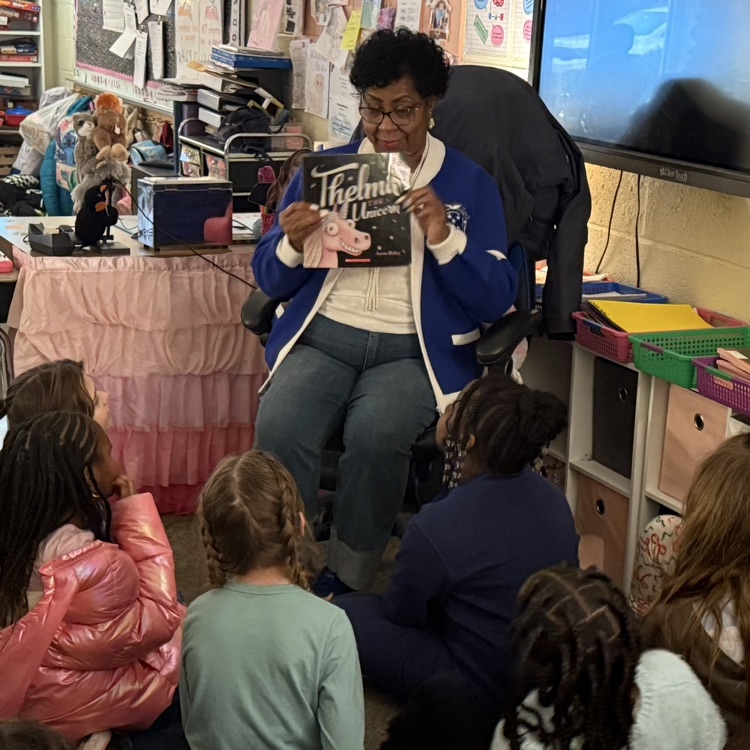 woman in blue sweater reading a book to students in a classroom