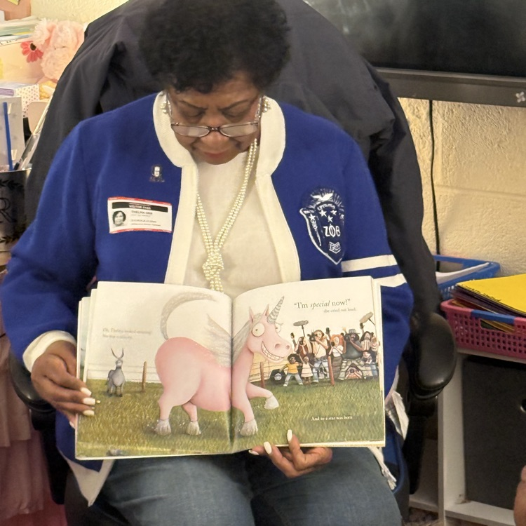 woman in blue sweater reading a book to students in a classroom