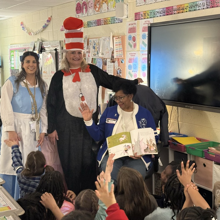woman in blue sweater reading a book to students in a classroom