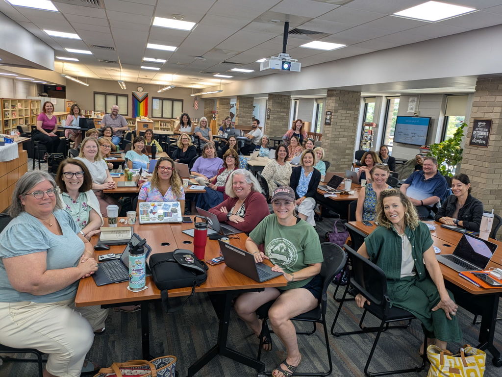 “A large group of school librarians smiling for a group photo at the end of the AI in the School Library forum. Participants are seated in clusters around tables with laptops, surrounded by bookshelves and natural light.”