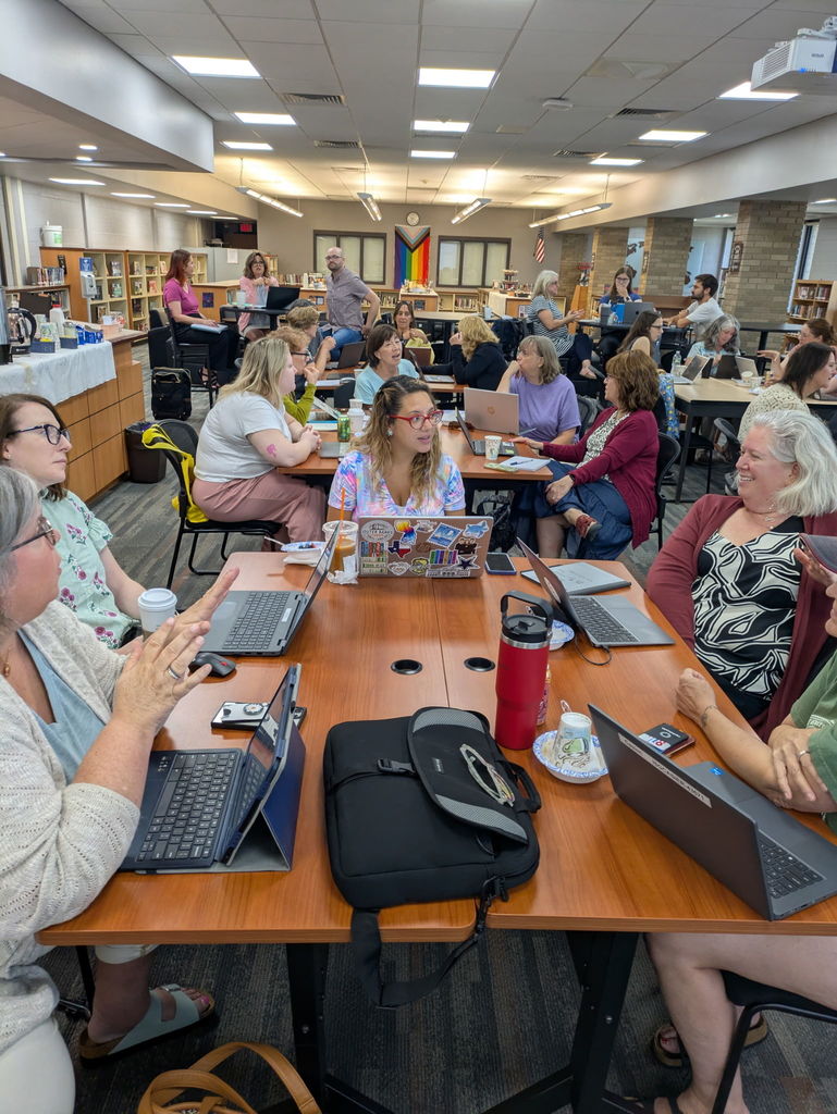 “Librarians actively engaged in small-group discussion around laptops during an AI workshop. Tables are covered with coffee cups, notebooks, and technology. The library space is bright and collaborative, with a pride flag and clock visible in the background.”