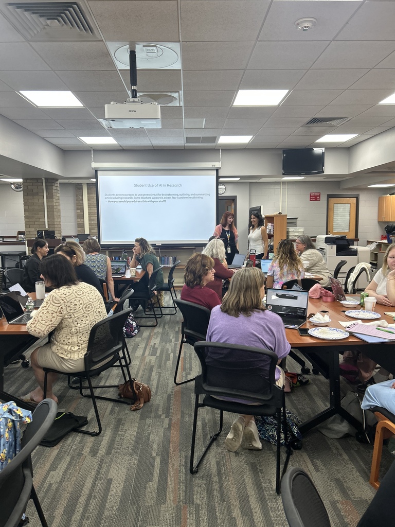 “School librarians seated in small groups at tables during a professional learning session in a school library. A projector displays a slide titled ‘Student Use of AI in Research’ as two facilitators stand at the front of the room.”