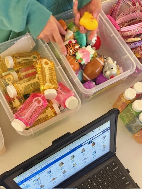 A close-up view captures a student browsing prizes at a school reward cart. The student's hands, wearing a light green sweatshirt, reach into a clear plastic storage bin filled with colorful small toys and novelty items. The student is holding a bright yellow squishy toy while sorting through other prizes that include animal-shaped figures, a small football-shaped toy, a zebra figure, a tiger-striped toy, pastel-colored fidget items, and assorted sensory toys in shades of purple, green, orange, and blue.  To the left sits another clear plastic bin filled with rectangular bottles containing gold-colored and pink-colored liquid. The bottles have white caps and textured sides that reflect the light, creating a sparkling appearance. Additional similar bottles are visible on the right side of the image, lined up on the table.  At the bottom of the image, an open laptop rests on the table. The laptop screen displays a digital behavior or reward tracking system with multiple student icons or avatars arranged in a gr