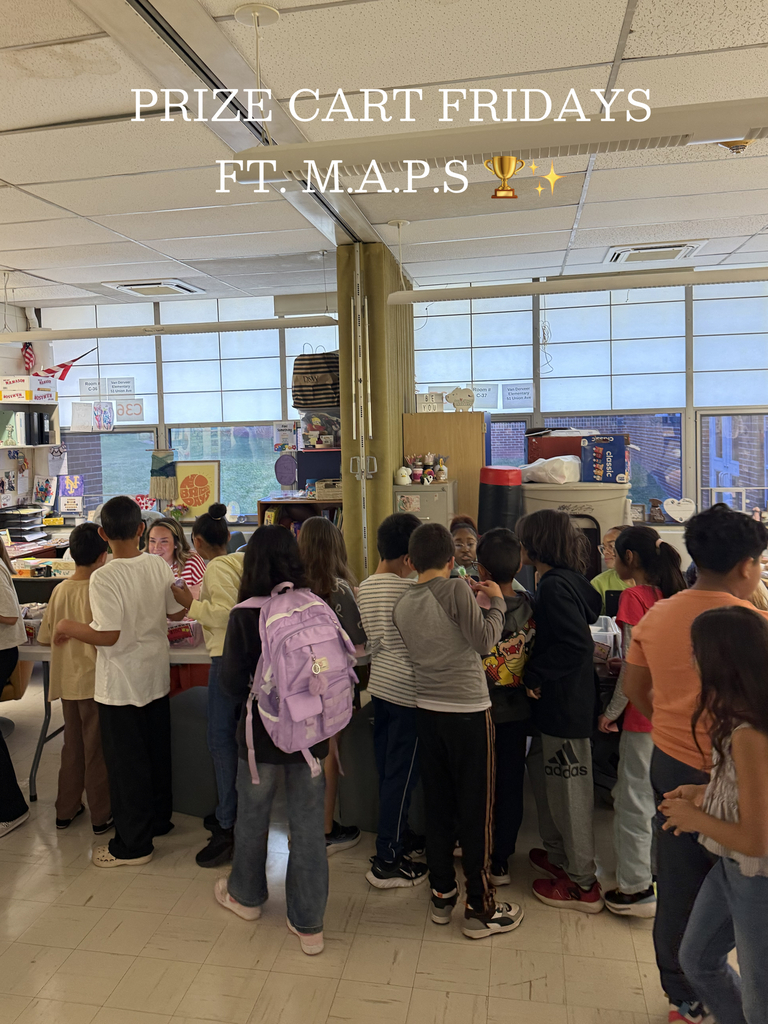 A large group of elementary-age students gather around several tables in a bright school classroom during a prize-selection event. White handwritten-style text overlaid across the top of the image reads, “PRIZE CART FRIDAYS FT. M.A.P.S.” Approximately twenty children stand closely together in a semicircle around tables that are mostly obscured by the crowd. One student near the center wears a light purple backpack, making the backpack stand out against the darker clothing around it.  The classroom has tall windows stretching across the back wall, allowing daylight to fill the room. Outside the windows, brick buildings and a grassy area are visible. Classroom furniture, shelves, artwork, and supplies line the walls. A teacher or staff member is partially visible behind the tables, interacting with the students. Decorative items, signs, and student projects are displayed throughout the room, creating a busy and welcoming school environment.  The room features a suspended ceiling with fluorescent light fixtures.