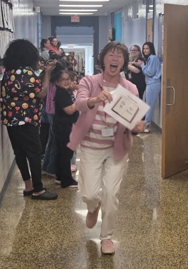 A woman joyfully running through a hallway, holding a certificate, while others line up behind her.