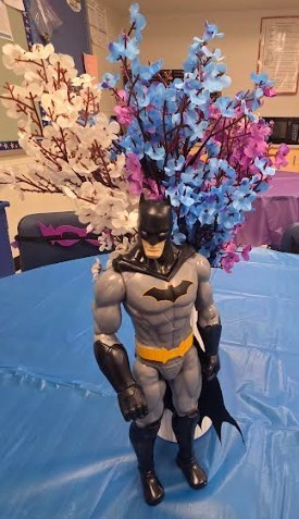 A Batman action figure stands in a vase filled with blue and pink flowers on a blue table.