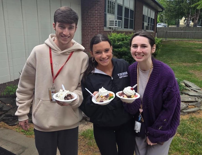 Three individuals stand outdoors holding bowls of ice cream, smiling. A house with brick walls and an air conditioner is behind them.