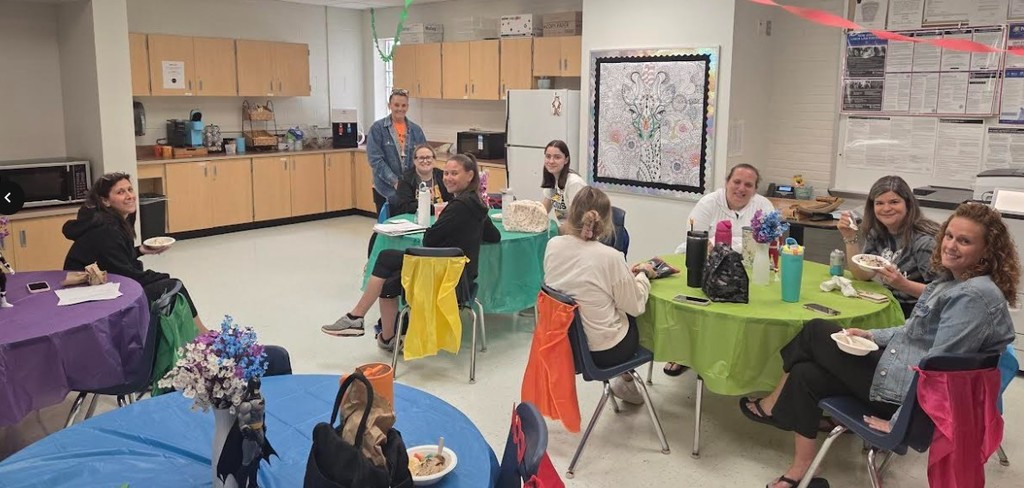 Group of adults sit around tables in a kitchen. Tables have bright tablecloths. One person stands by a wall.