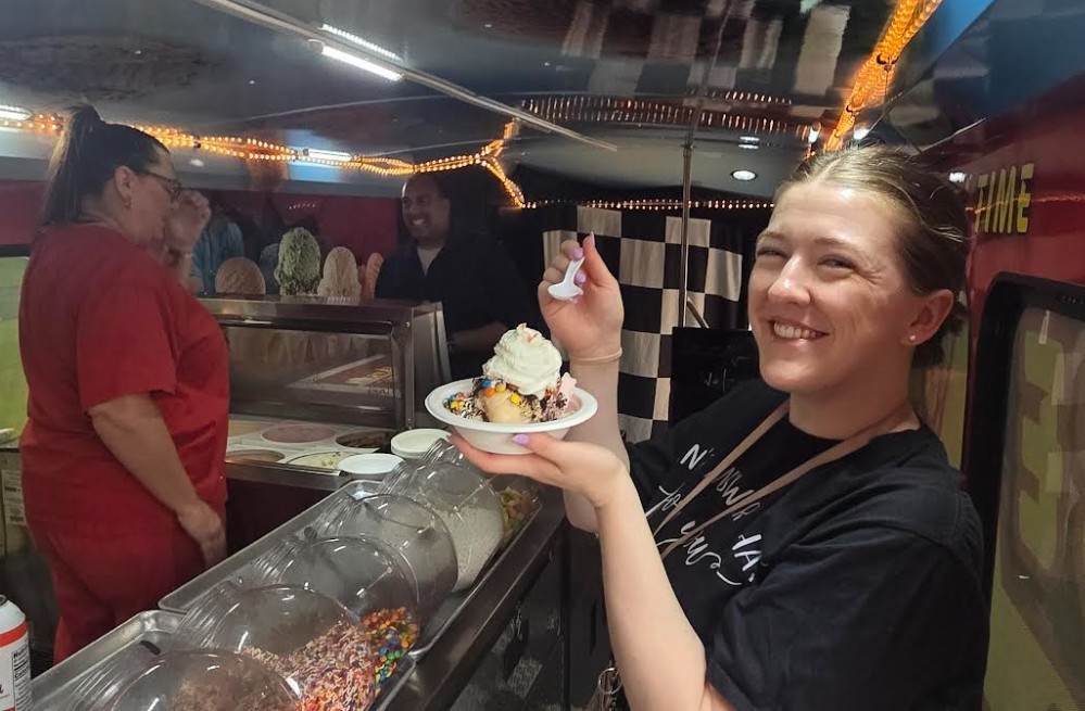 A woman holds a dessert on a plate. Another woman in red stands behind the counter.