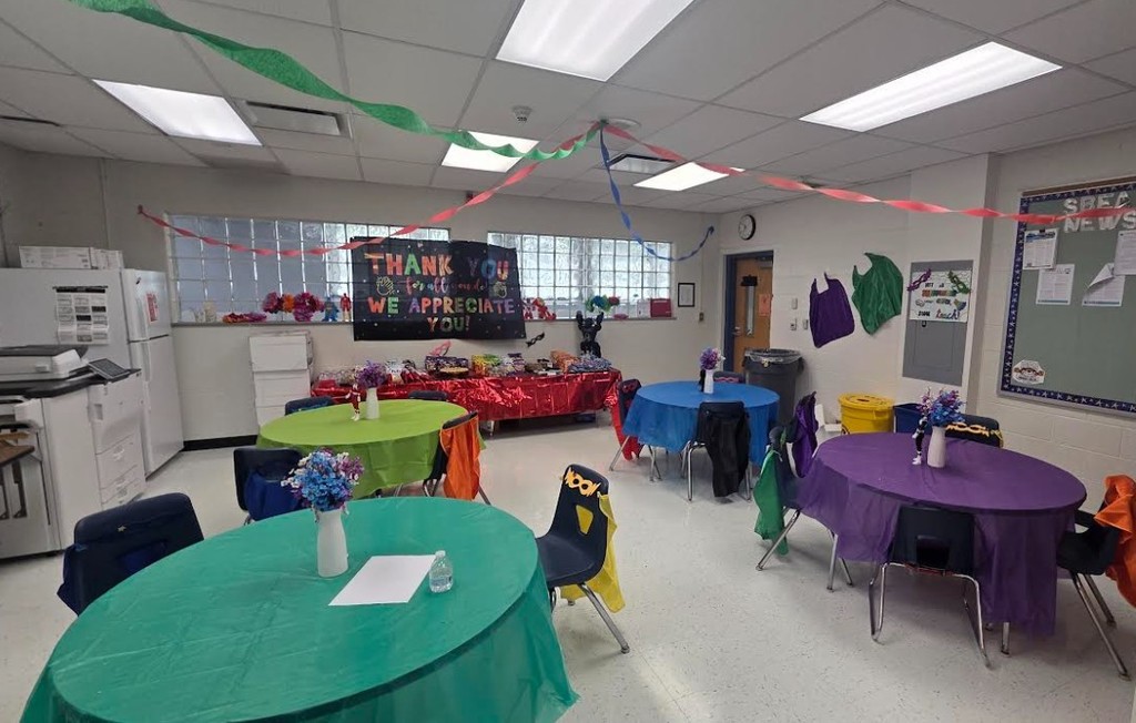 A classroom with various colored tables and chairs, a white refrigerator, and a bulletin board.