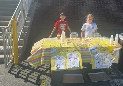 Two kids standing behind a table covered with a yellow and white striped tablecloth and holding plastic cups.