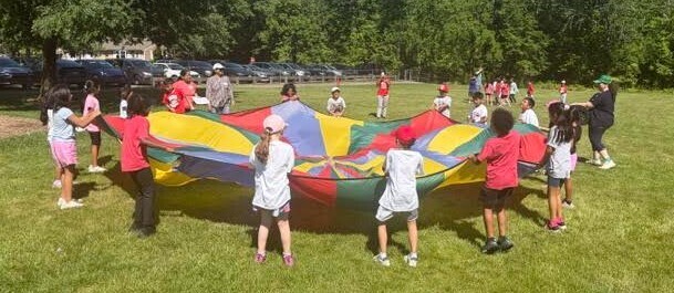A group of children playing on a colorful, large bouncy mat in a park with trees.