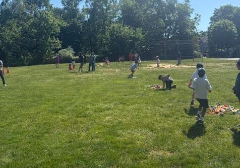 A group of people of various ages playing in a grassy field with trees in the background.