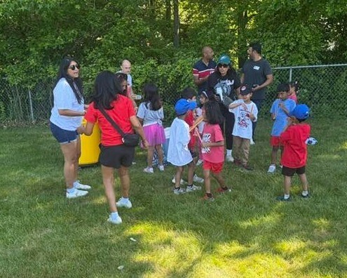 Group of adults and children standing on grassy field with trees and fence. Some children wear hats.